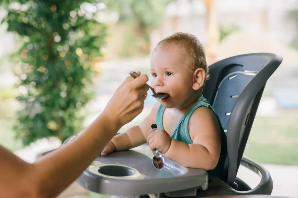 Parent spoon feeding baby in a high chair, from our baby food chart by month feeding guide