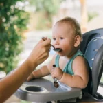 Parent spoon feeding baby in a high chair, from our baby food chart by month feeding guide